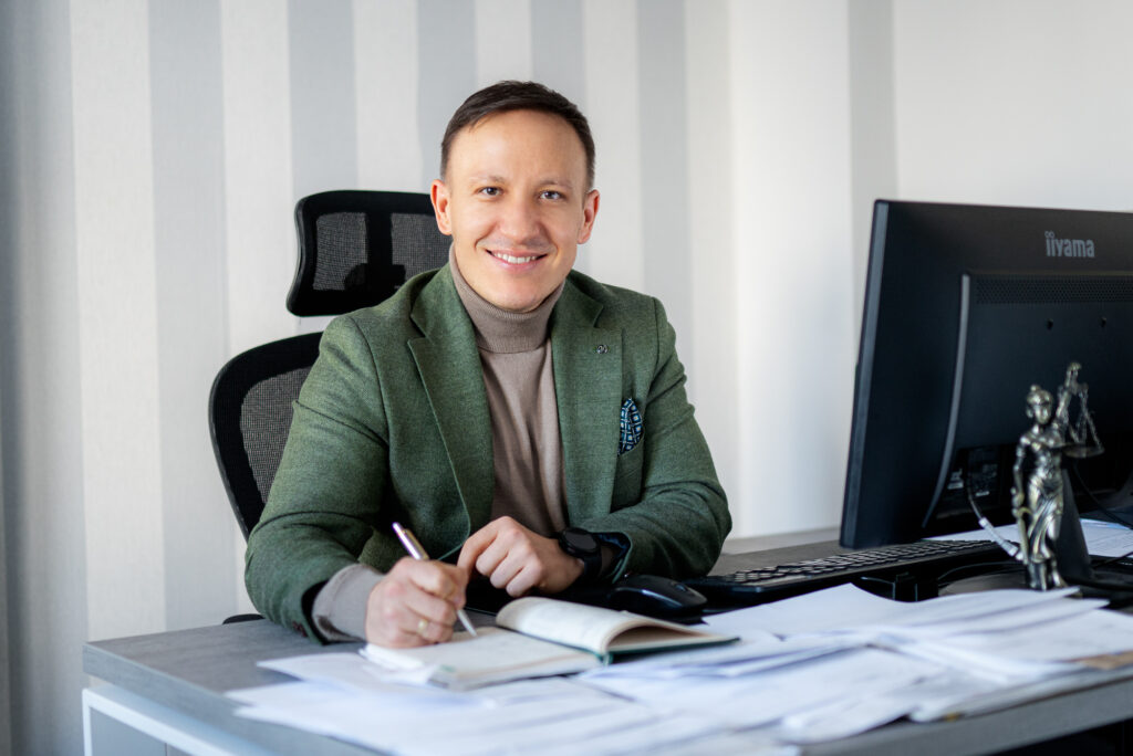 Uśmiechnięty mężczyzna w marynarce siedzący przy biurku i podpisujący dokumenty w biurze prawnym